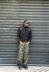 Sort polstret vest over en sort langærmet skjorte, camouflage løstsiddende bukser, sorte støvler og solbriller; lænet op ad en bølgepladevæg.