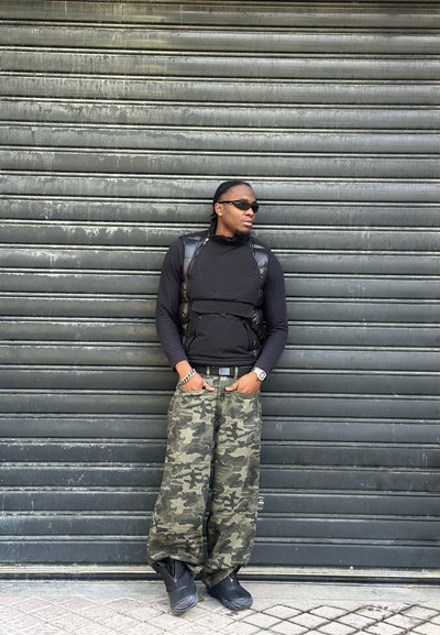 Chaleco acolchado negro sobre una camisa de manga larga negra, pantalones holgados de camuflaje, botas negras y gafas de sol; apoyado contra un fondo de metal corrugado.