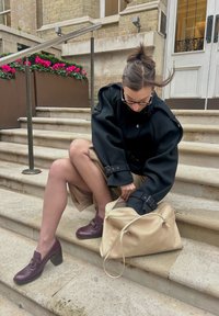 Svart oversized kåpe, beige skinnveske, lilla blokkhælte sko; modellen sitter på steintrapper med rosa blomsterpotter i bakgrunnen.