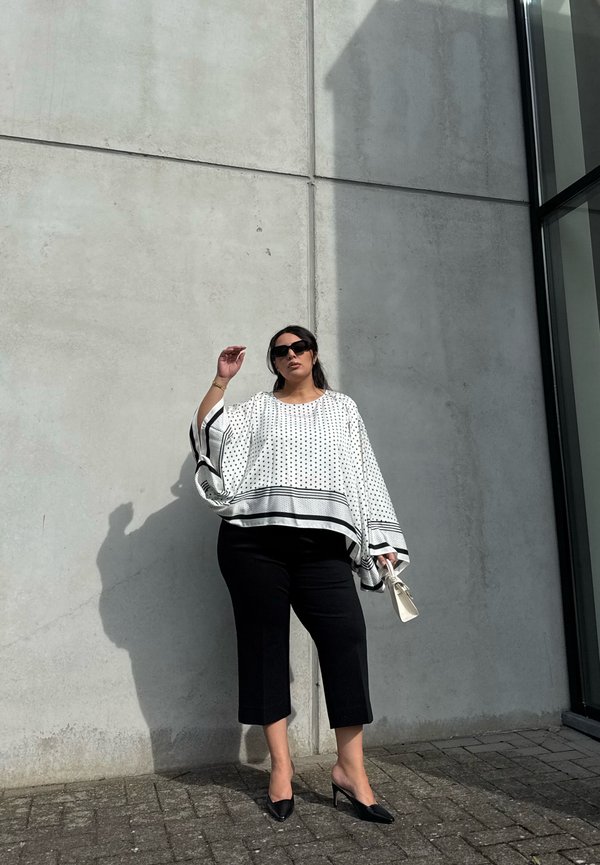 Mujer con pantalones negros cortos y una blusa blanca con lunares, está apoyada contra una pared de hormigón, sosteniendo un pequeño bolso blanco.