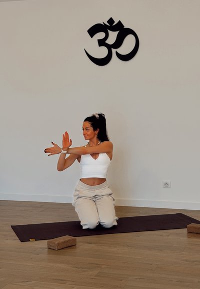 Práctica de yoga sobre una esterilla morada con una persona arrodillada vestida con ropa clara, demostrando una postura con bloques de madera cerca.