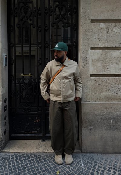 Hombre con chaqueta beige, pantalones oliva, gorra de béisbol verde y bolso al hombro, de pie mirando hacia la izquierda junto a una puerta de hierro forjado negra en una calle pavimentada con piedra.