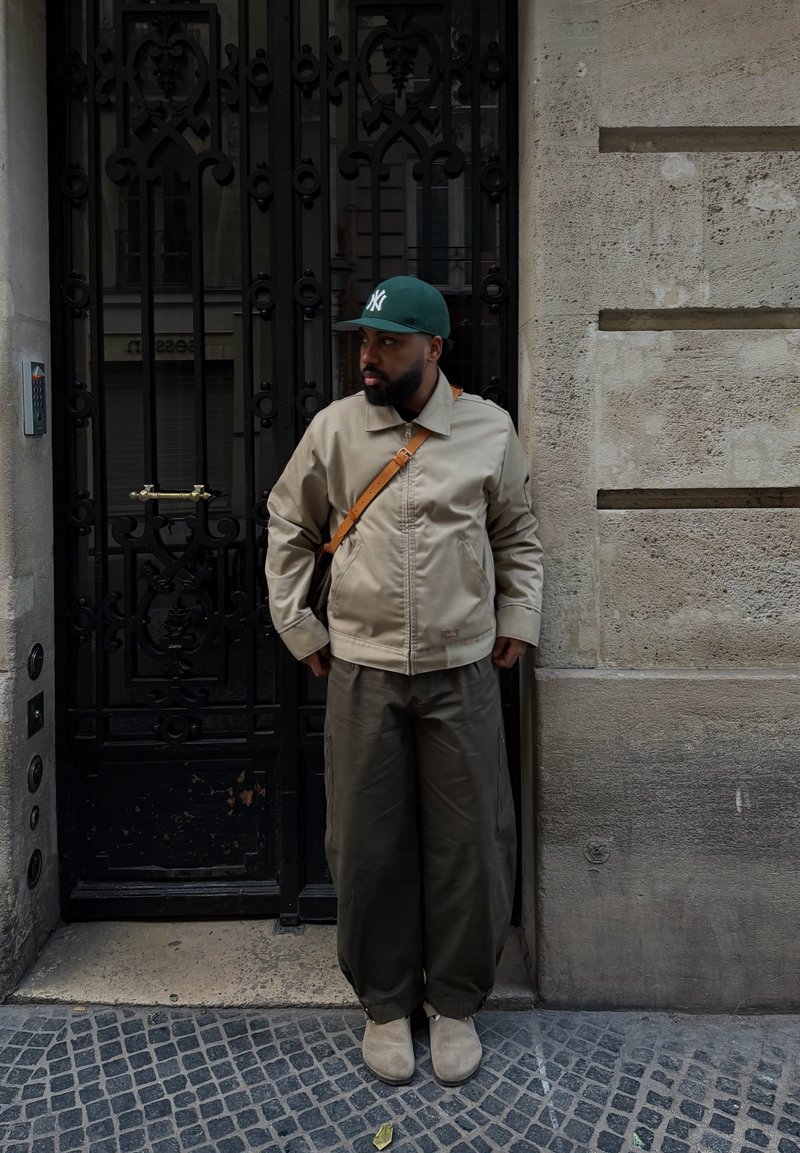 Homme en veste beige, pantalon olive, casquette de baseball verte et sac à bandoulière, debout face à gauche près d'une grille en fer forgé noire sur une rue pavée de pierres.