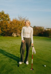 Weißes Langarmshirt mit halbem Reißverschluss, kombiniert mit lockeren olivgrünen Hosen. Steht auf einem Golfplatz, hält einen Putter und ein Handtuch in der Nähe eines Lochs.