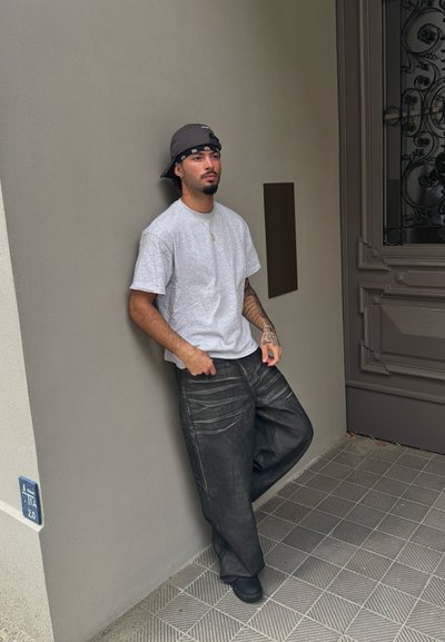 Camiseta de algodón gris, pantalones de mezclilla negros holgados con detalles de costura visibles y un sombrero de cubo oscuro. Persona apoyada contra una pared.