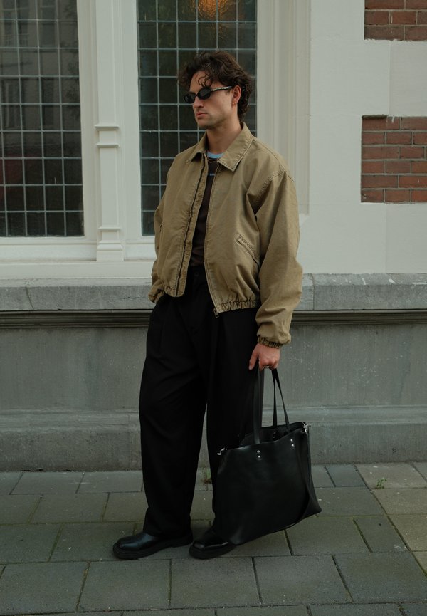Hombre con gafas de sol, chaqueta beige, pantalones negros y zapatos negros sosteniendo una gran bolsa negra de pie en una acera de la ciudad cerca de una ventana.