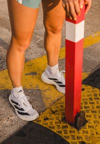 Witte sportschoenen met zwarte strepen, gedragen met witte sokken, staande op een gestructureerd gele ondergrond nabij een rood-witte paal.