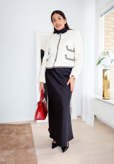 Mujer con chaqueta blanca con botones y falda larga negra, sosteniendo un bolso rojo, de pie en el interior cerca de un espejo de cuerpo entero y una ventana.