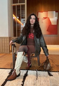 Giubbotto di pelle marrone su un vestito aderente marrone con una grafica rossa. Stivaletti da cowboy beige con dettagli marroni. Collant texturizzati con dettagli laterali.