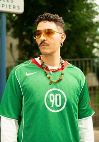 Maillot Nike vert avec des détails blancs et un logo circulaire "90". Porté par-dessus une chemise à manches longues blanche, accessoirisé avec des colliers de perles colorées.