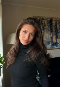 Młoda kobieta o długich brązowych włosach, ubrana w czarny golf, stoi w pomieszczeniu przy oknie, w tle lampa i abstrakcyjny obraz.