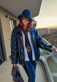 Chaqueta de cuero negra sobre una camisa blanca, combinada con un cinturón negro con hebilla plateada, pantalones negros, un sombrero de fieltro negro y gafas de sol.