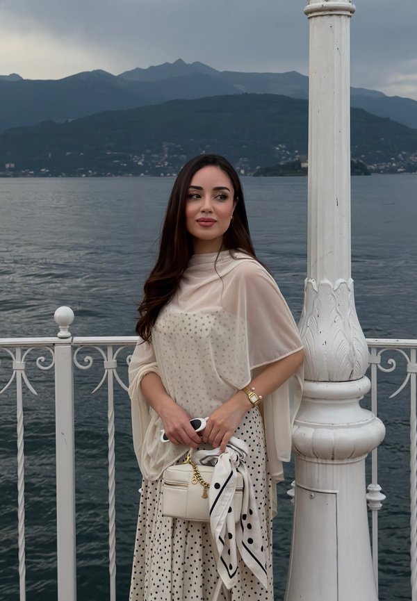 Jeune femme en robe à pois et châle se tient près d'une rambarde blanche avec un sac à main, surplombant un lac et des montagnes sous un ciel nuageux.