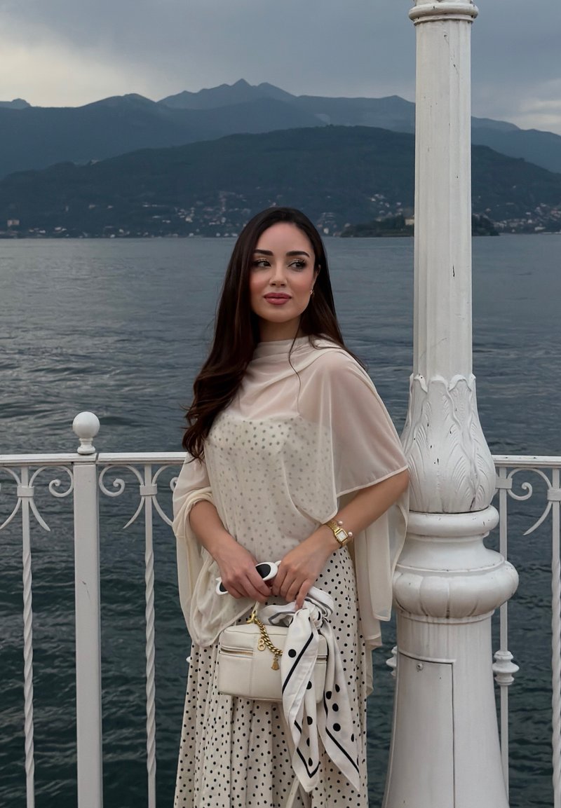 Jeune femme en robe à pois et châle se tient près d'une rambarde blanche avec un sac à main, surplombant un lac et des montagnes sous un ciel nuageux.