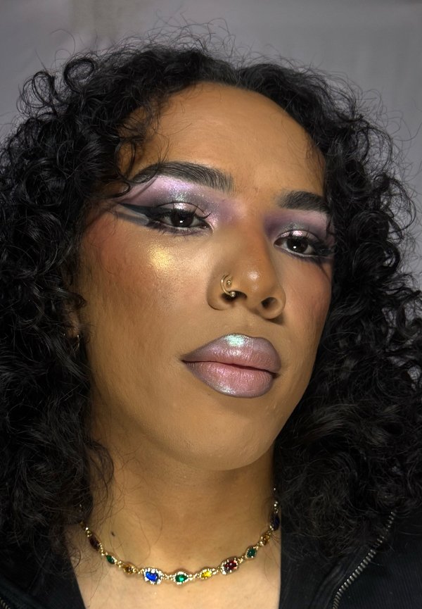 Face with curly hair, bold purple and black eye makeup, metallic lips, nose piercing, and multicolored gem necklace against gray background.