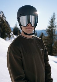 Casque de ski noir avec lunettes de ski miroir, porté sur un t-shirt à manches longues foncé. Paysage enneigé et arbres à feuilles persistantes en arrière-plan.
