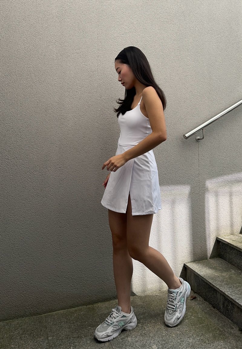 Weißes ärmelloses Kleid mit seitlichem Schlitz, kombiniert mit grauen und weißen Sportschuhen. Der Hintergrund zeigt eine strukturierte graue Wand und Treppen.