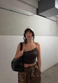 Jonge vrouw in een witte pet, een donker geraffineerde crop-top en camouflagebroek staat in een betonnen gang met een zwarte schoudertas.