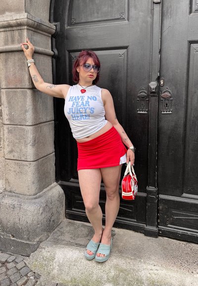 La modelo lleva una camiseta blanca con texto azul, una mini falda roja y sandalias de plataforma aqua, sosteniendo un pequeño bolso rojo junto a una puerta negra.