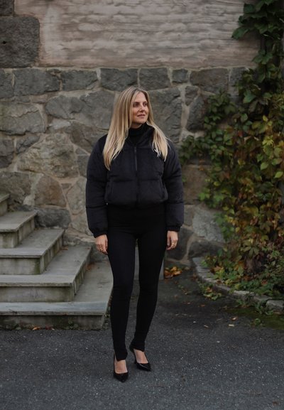 Chaqueta puffer negra con cierre de cremallera, combinada con leggings negros y tacones. El tejido parece suave, con una silueta ajustada.