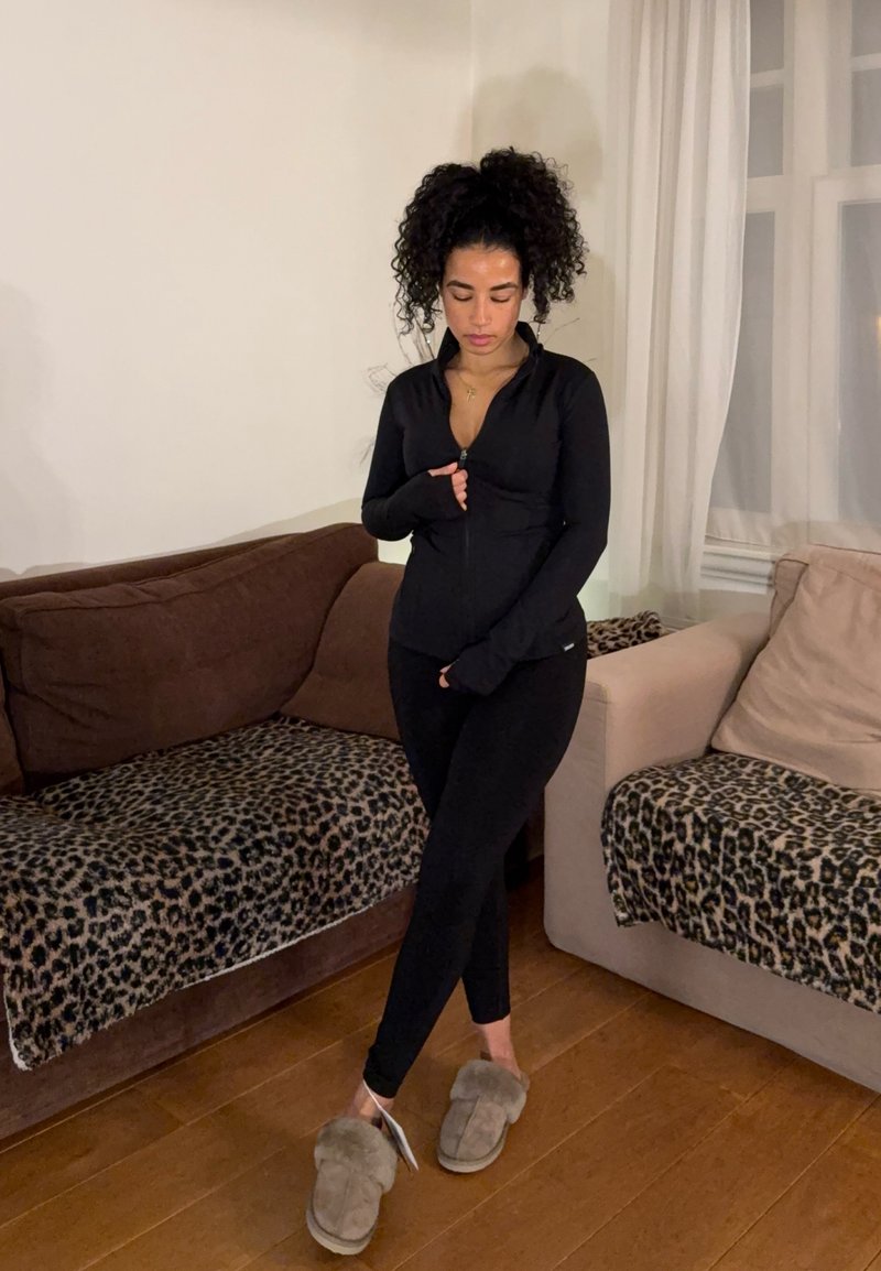 Femme aux cheveux bouclés portant des vêtements de sport noirs et des chaussons duveteux, debout entre deux canapés recouverts de plaids à imprimé léopard dans un salon.