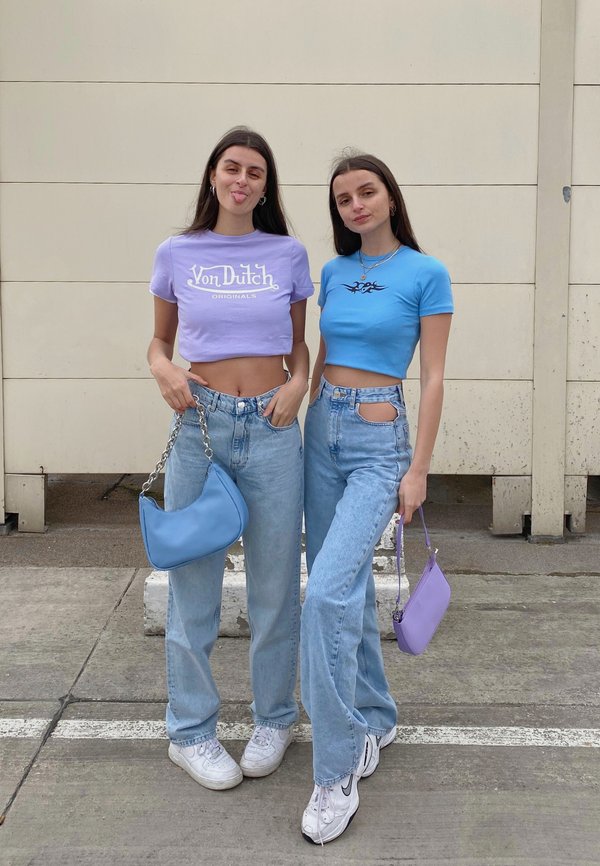 Dos mujeres están de pie una al lado de la otra, llevan tops cortos: uno es de color lavanda con texto blanco y el otro azul con un diseño negro. Ambas visten jeans de un azul claro y sostienen pequeñas bolsas.