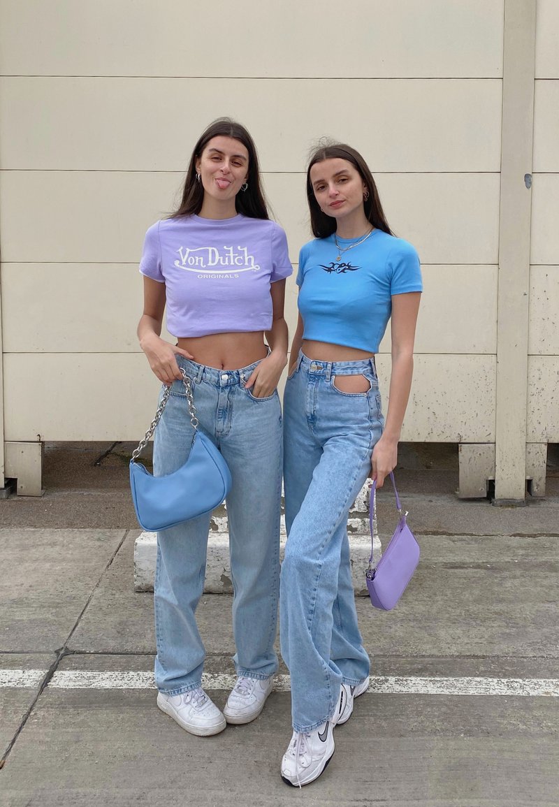 To kvinder står side om side iført cropped tops: en lavendel med hvid tekst og en blå med et sort design. Begge har lyseblå jeans på og holder små tasker.