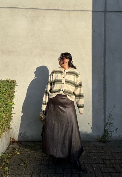 Cárdigan de punto con rayas verdes y crema, falda maxi de cuero marrón, accessorios con gafas de sol y un bolso pequeño, de pie contra una pared.