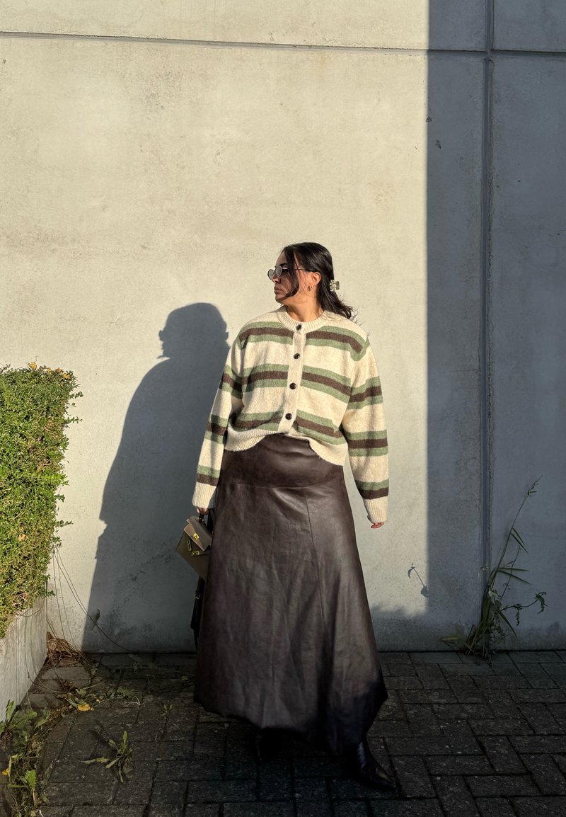 Pletena kardigan s zelenim i kremastim prugama, smeštanje smeđe kožne maxi suknje, s dodacima poput sunčanih naočala i male torbice, stoji pored zida.