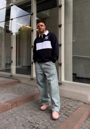 Chemise à rayures noires et blanches à manches longues, pantalon large vert clair et chaussures roses. Debout sur des pavés à côté d'un bâtiment en verre.