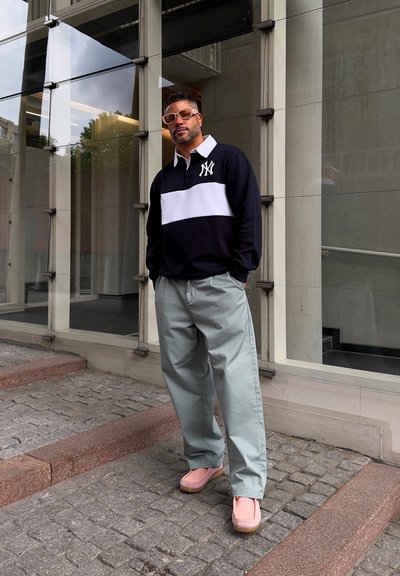 Camisa de rayas blancas y negras de manga larga, pantalones de pierna ancha verde claro y zapatos rosas. De pie sobre adoquines al lado de un edificio de cristal.