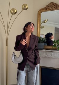 Blazer marron foncé avec un col en V profond, fermeture à boutons et détail de laçage sur le côté. Sac à main argenté à finition lisse, assorti à un pantalon gris clair.