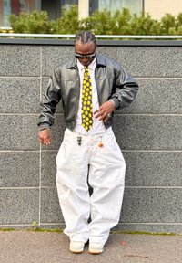 Casaco de couro preto, camisa branca com riscas, gravata com rosto sorridente, calças brancas largas, porta-chaves decorativos e ténis brancos com detalhes verdes.