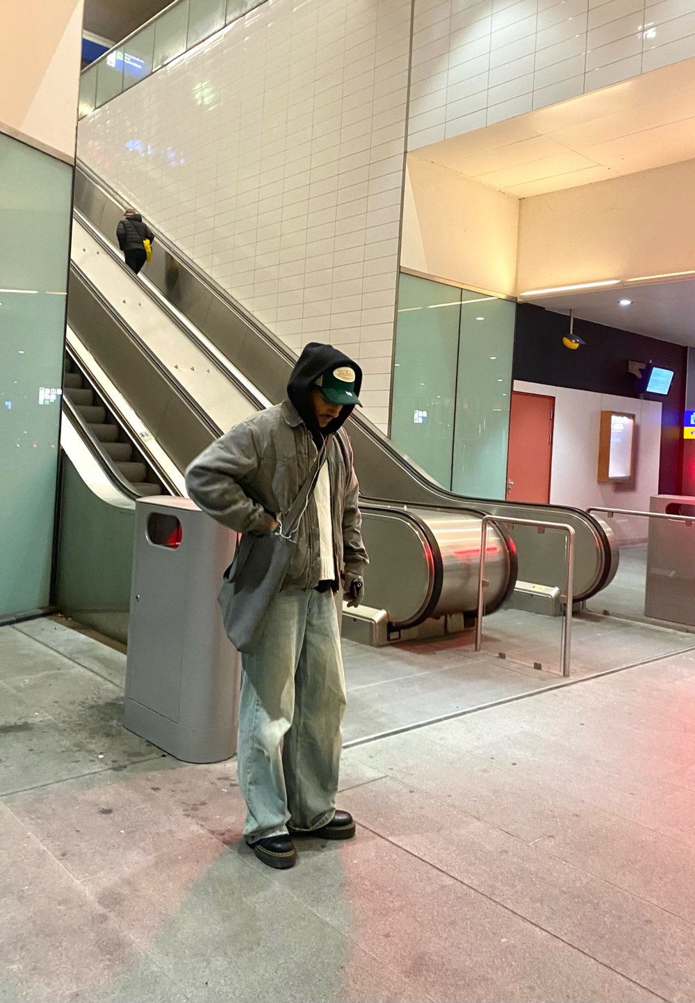 Una persona con una giacca oversized grigia, jeans larghi e scarpe nere si trova vicino a una scala mobile in uno spazio moderno ben illuminato.