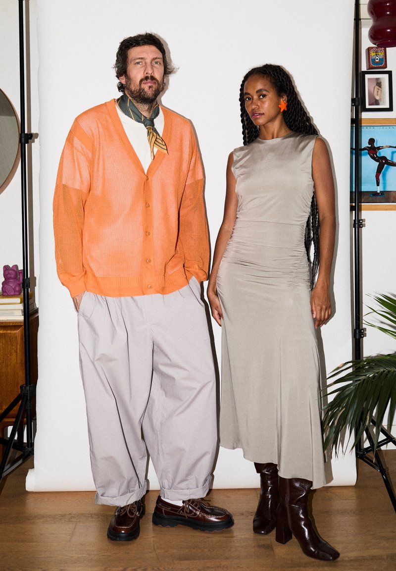 Άνδρας με πορτοκαλί ζακέτα και φαρδιά παντελόνια στέκεται δίπλα σε γυναίκα με μπεζ φόρεμα χωρίς μανίκια και καφέ μπότες σε λευκό φόντο.