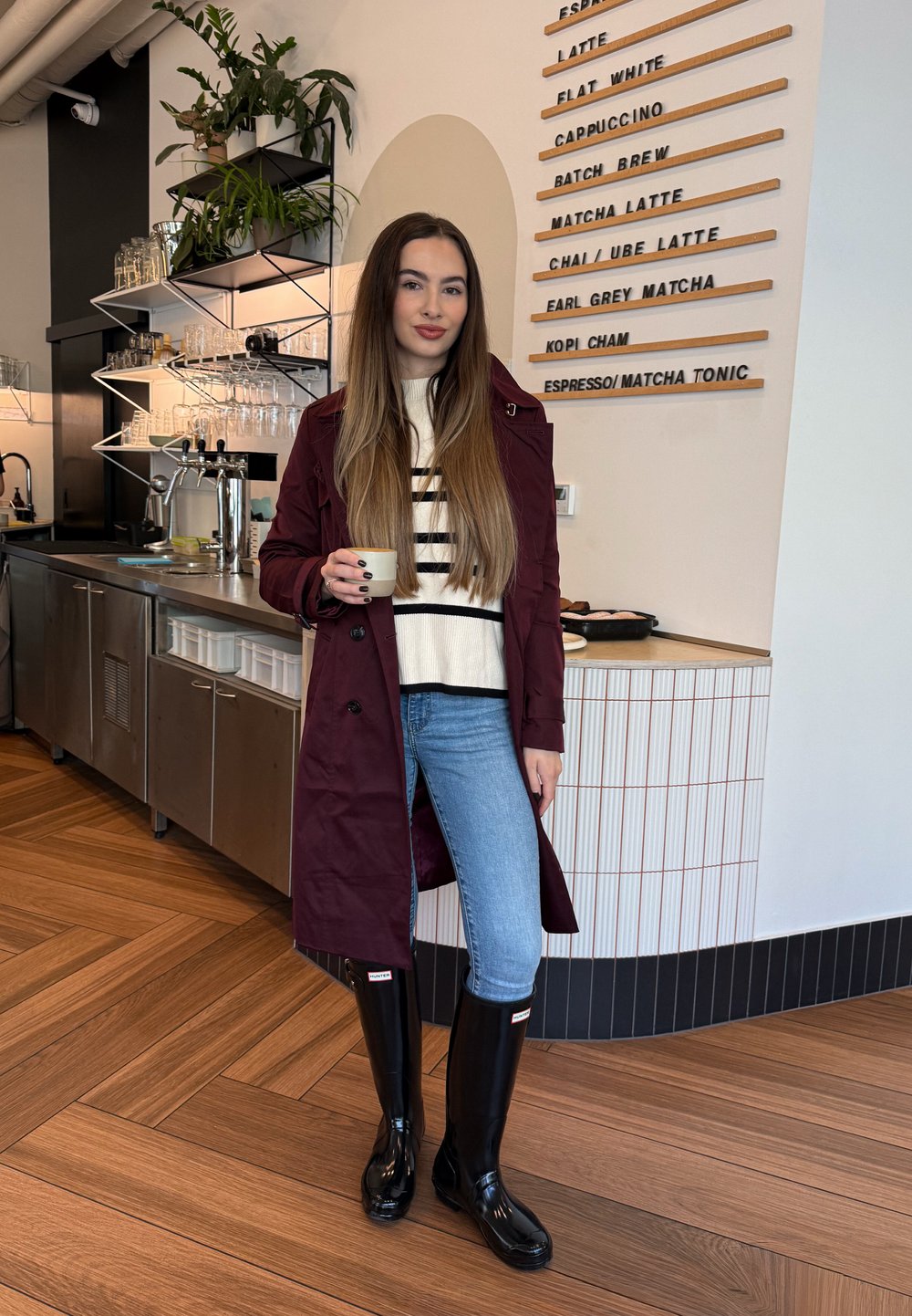 Kvinne i maroon frakk, stripete genser, jeans og svarte støvler som holder en kopp inne i en kafé med en kaffemeny på veggen.