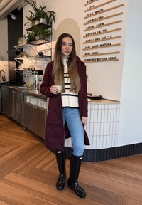 Kvinne i maroon frakk, stripete genser, jeans og svarte støvler som holder en kopp inne i en kafé med en kaffemeny på veggen.