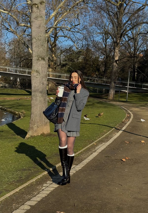 Ung kvinde i grå blazer, ternet nederdel og støvler holder en kop kaffe og står på en stis ved en park nær en dam med ænder og træer uden blade.