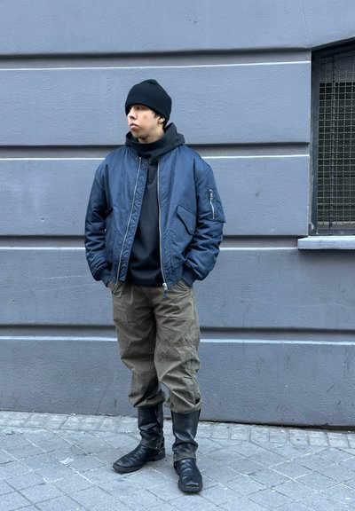 Chaqueta bomber marina, sudadera negra con capucha, pantalones cargo oliva y botas negras hasta la rodilla. De pie contra una pared gris con una ventana.
