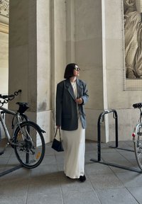 Grijze oversized blazer, witte maxi jurk, zwarte handtas, donkere schoenen, staand naast fietsen in een doorgang met getextureerde stenen muren.