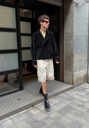 Joven con gafas de sol, chaqueta negra, pantalones cortos beige y zapatillas negras saliendo de un edificio hacia una acera con baldosas.