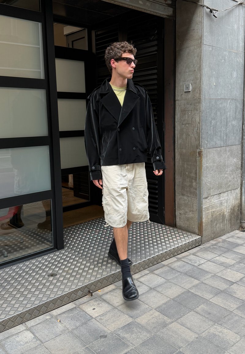 Jeune homme portant des lunettes de soleil, une veste noire, un short beige et des chaussures noires, sortant d’un bâtiment sur un trottoir carrelé.