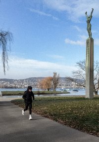 Trkač u crnoj jakni i hlačama na popločanoj stazi blizu obale, s zelenim kipom na visokom kamenom postamentu u pozadini.