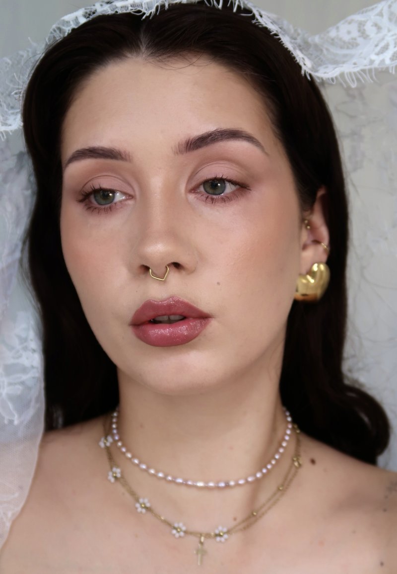 Jeune femme aux cheveux foncés porte un anneau nasal en septum doré, une grande boucle d'oreille en forme de cœur en or, des colliers superposés de perles et de fleurs, et un voile en dentelle blanche.