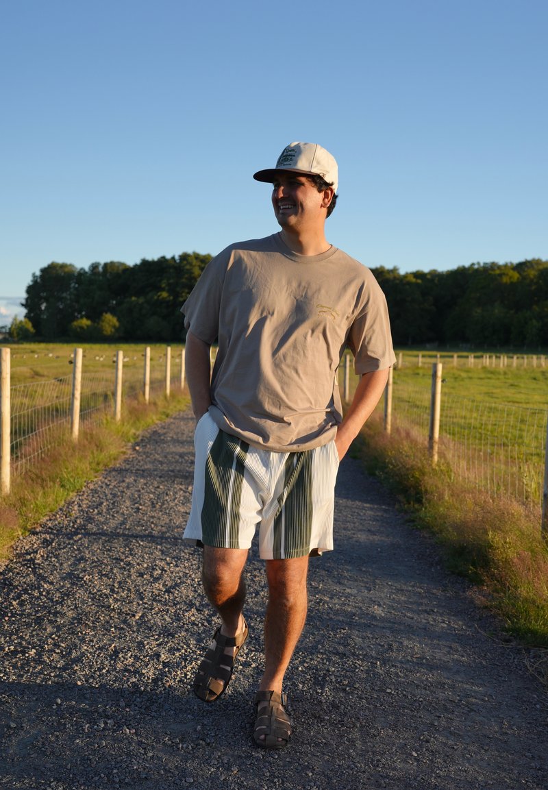 Homme portant une casquette, un t-shirt et un short rayé marchant sur un chemin de gravier entre des champs herbeux clôturés au coucher du soleil.