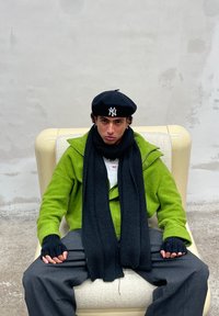 Πράσινο fleece jacket, μαύρο κασκόλ, γάντια χωρίς δάχτυλα και μαύρη μπερέ. Το άτομο κάθεται σε μια μπεζ καρέκλα μπροστά σε έναν γκρι τοίχο.