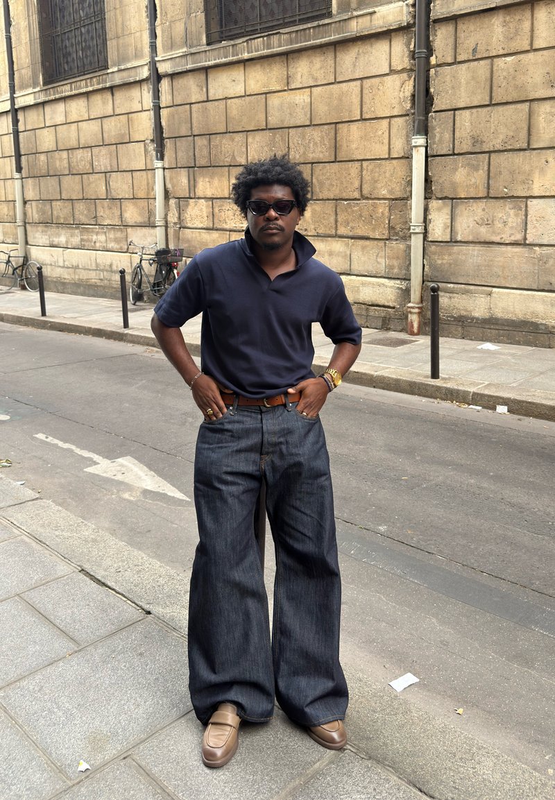 Homme portant des lunettes de soleil, un polo bleu marine, un jean large foncé et des chaussures beige, debout dans une rue de la ville à côté d'un bâtiment en pierre.