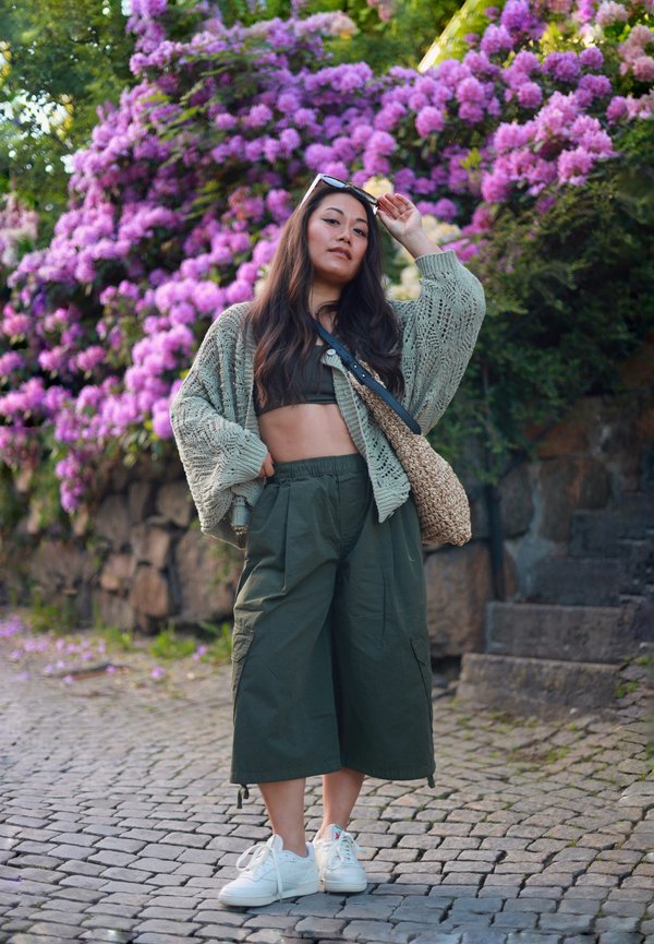 Jeune femme en pantalon court vert et cardigan pose sur un chemin pavé avec des fleurs violettes et des marches en pierre derrière elle.