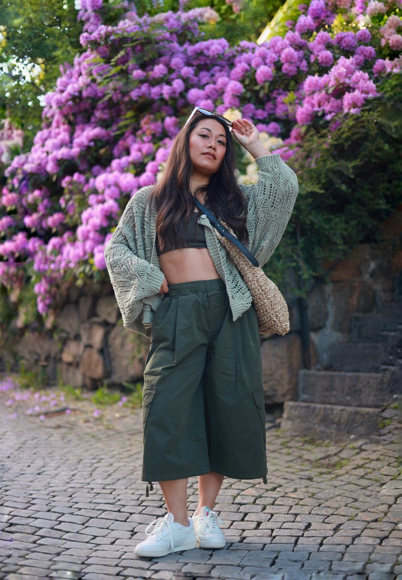 Young woman in green cropped trousers and cardigan poses on cobblestone path with purple flowers and stone steps behind her.
