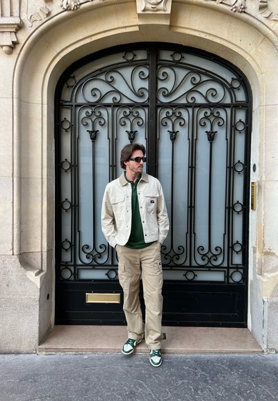 Chaqueta de mezclilla beige sobre un polo verde, pantalones cargo caqui y zapatillas verdes y blancas, de pie frente a una puerta decorativa de metal negro.